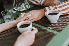 Close up of young man with the ring on his finger taking hand of his girlfriend. They sitting at the table with white cups of coffee