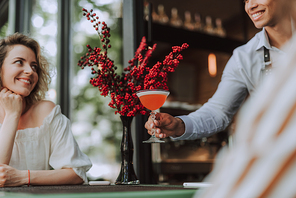 Beautiful young woman looking at barman with smile while he placing glass of cold drink on the table. Vase with bouquet of red berries on background