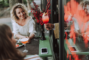 Beautiful young woman looking at glass of cocktail in bartender hand and smiling. She sitting at the table with modern smartphones and bouquet of red berries