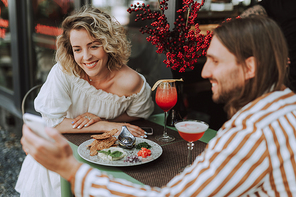 Bearded man holding smartphone while charming lady looking at display and smiling. They sitting at the table with food and cocktails