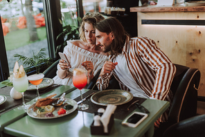 Charming lady and bearded man watching video on smartphone. They sitting at the table with food and cocktails