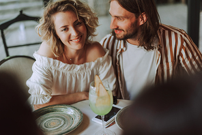 Close up portrait of bearded man looking at his charming girlfriends and smiling. They sitting at the table next to each other