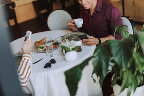 Close up of smiling young man with cup of hot drink and his friend checking messages on smartphones. They sitting at the table with food and beverages