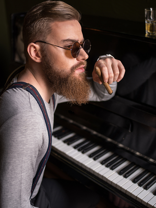 Portrait of cheerful bearded musician smoking a cigar. He is sitting near the piano and looking aside pensively. The man has retro style