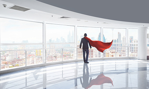 Businessman in a suit and hero cape and looking at the city, stands in empty office. Victory concept, morning scene .
