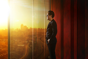 Young asian businessman looking out over the city through office transparent glass window . Sunrise scene .