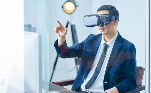 Asian businessman wearing virtual reality goggle and pointing up in a office .