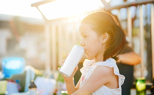 Portrait of a happy asian little girl drinking a soda refreshment from a can in the park during sunset