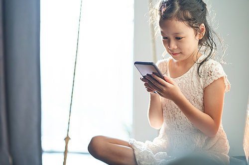 Pretty happy asian little girl  holding smartphone sitting on living room sofa and feeling happy.