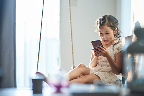 Happy cheerful little girl holding smartphone sitting on living room sofa and feeling surprised .