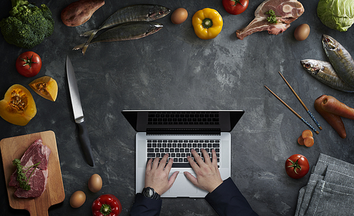 Man in the kitchen searching for recipes on his laptop with fresh ingredients on cement kitchen worktop, top view