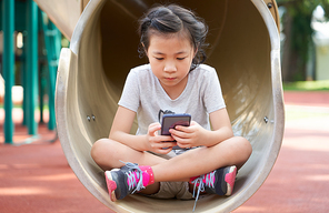 Little girl sitting in the park playing with smartphone phone, kids mobile phone addiction concept.
