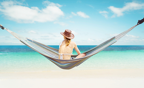Woman in hat sitting on hammock on the beach and enjoying the beautiful seascape .