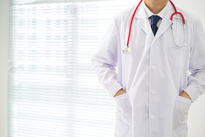 Unknown doctor posing two hand in pocket in the office, he is wearing a stethoscope .