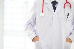 Unknown doctor posing two hand in pocket in the office, he is wearing a stethoscope .