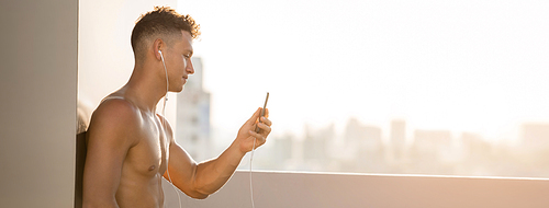 Banner images of shirtless handsome sports man wearing earphones listening to music from mobile phone relaxing after workout on rooftop in evening sunlight
