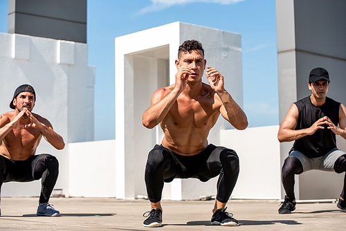 Group of fit sports men doing squat bodyweight workout training outdoors on building rooftop in sunlight - exercisse in the open air concept