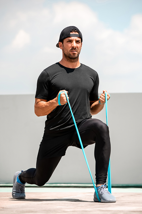 Handsome Latino sports man doing lunge workout with resitance band outdoors in the sun