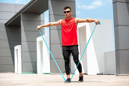 Handsome sportsman doing shoulder lateral raise exercise with resistance band outdoors on rooftop in the sun