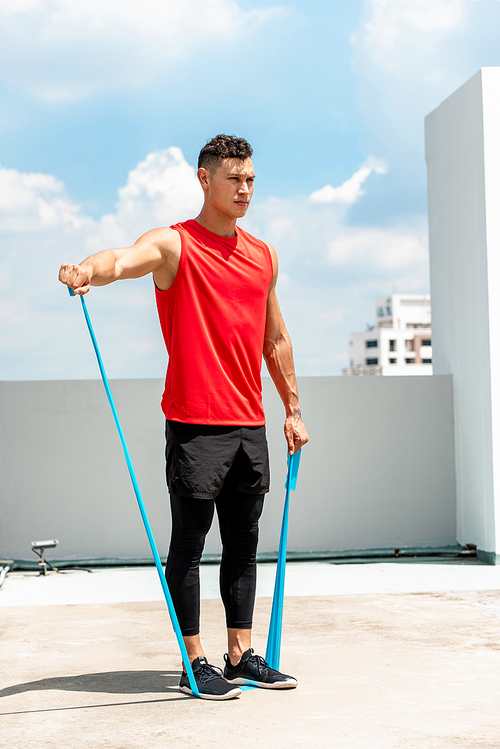 Handsome sports man doing shoulder lateral raise exercise with resistance band outdoors on rooftop in the sun