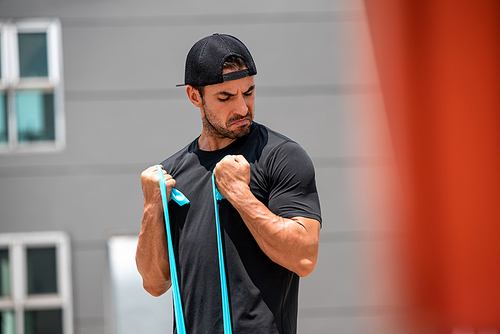 Fit muscular Latino sports man doing bicep curl exercise with resistance band outdoors at home in sunlight