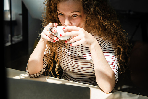 Young Caucasian woman drinking coffee while watching video on laptop computer at home in early morning