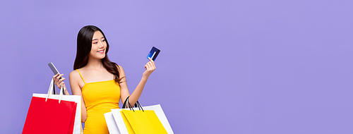 Portrait of young smiling beautiful Asian woman making online payment through phone via credit card while carrying colorful shopping bags in isolated purple banner background