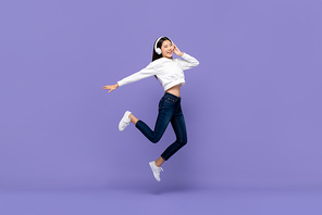 Happy young Asian woman jumping and listening to music on headphones isolated on purple background