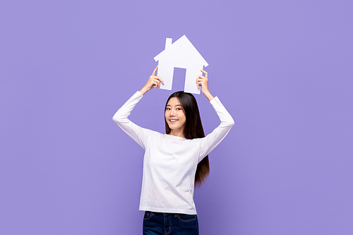 Smiling beautiful Asian woman holding house symbol overhead isolated on purple background
