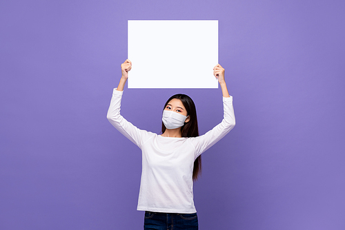 Young Asian woman wearing medical mask holding white paper board with empty space for text overhead