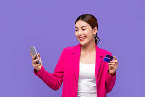 Happy beautiful young Asian woman in pink blazer shopping with credit card via mobile phone isolated on purple background