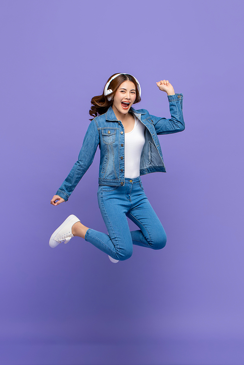Happy young Asian woman jumping and listening to music on headphones isolated on purple studio background