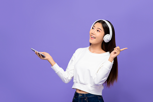 Portrait of young smiling Asian woman listening to music on mobile phone using wireless headphone in purple isolated studio background