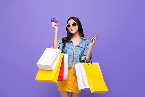 Happy young Asian girl carrying colorful shopping bags with credit card and mobile phone in hands isolated in purple background
