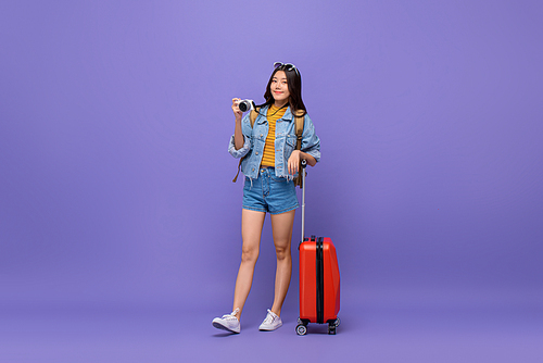Young smiling Asian tourist girl in casual attire with camera and baggage isolated on colorful purple background