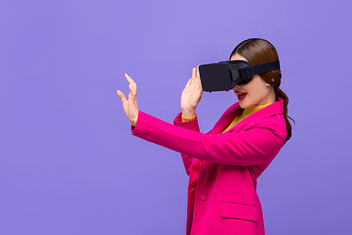 Young woman wearing virtual reality or VR glasses reaching hand out to touch something on purple isolated background