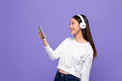 Portrait of young smiling Asian woman listening to music on mobile phone using wireless headphone in purple isolated studio background