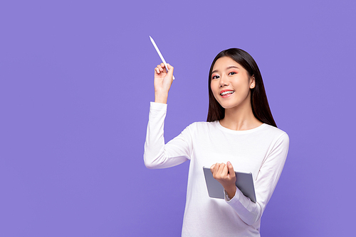 Smiling beautiful Asian woman in white attire pointing with stylus to copy space and holding digital tablet isolated on purple background