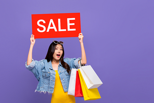 Surprised fashionable Asian woman carrying colorful shopping bags raising red sale sign isolated on purple background