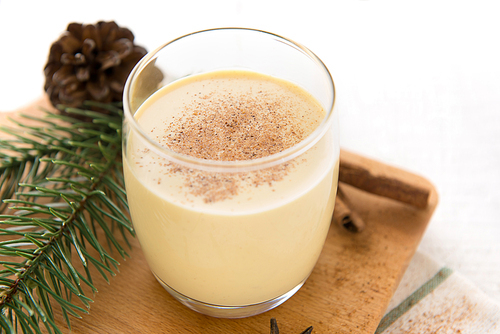 Spiked eggnog with ground nutmeg served in glass on wooden platter decorated with pine and cinnamon sticks on white table