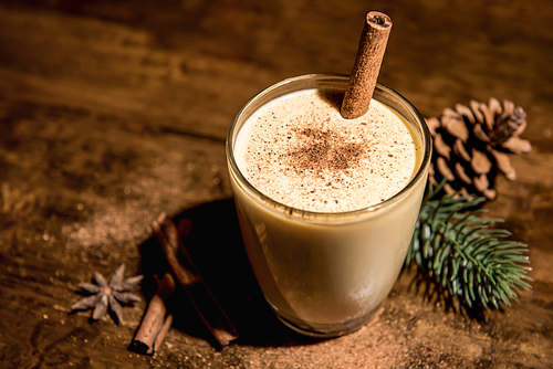 Homemade traditional Christmas eggnog drink in a glass with ground nutmeg, cinnamon and decorating items on wood table, preparing for celebrating festive holiday season