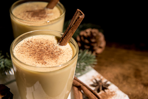 Homemade traditional Christmas eggnog drinks in glasses with ground nutmeg, cinnamon and decorating items on wood table, preparing for celebrating festive holiday season