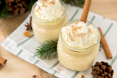 Traditional Christmas eggnog drinks with whipped cream, ground nutmeg, cinnamon and decorating items on wood table, preparing for celebrating festive holiday season