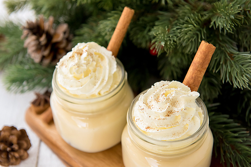 Traditional Christmas eggnog drinks with whipped cream, ground nutmeg, cinnamon and decorating items on wood table, preparing for celebrating festive holiday season