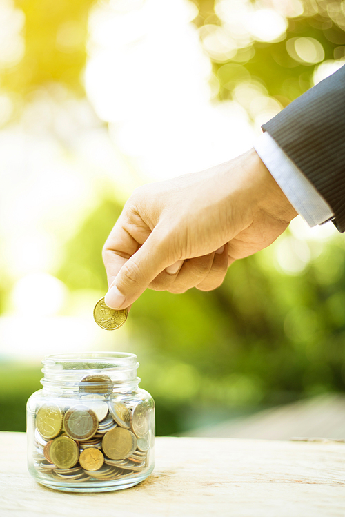 Businessman hand putting money (coin) into the glass jar - savings, investment and donation concepts