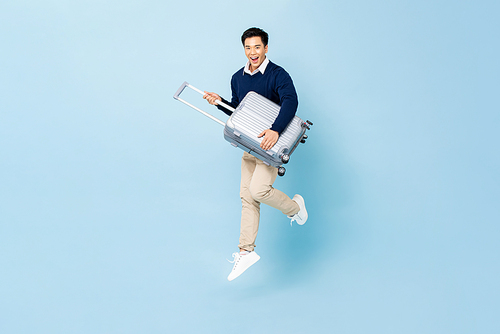 Jumping portrait of young happy smiling handsome Asian tourist man with baggage ready to travel isolated on light blue studio background