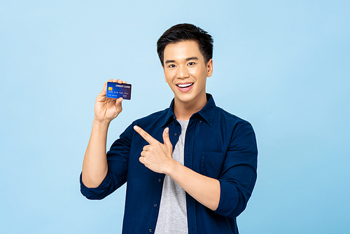 Happy smiling young handsome Asian man pointing to credit card in hand isolated on light blue background