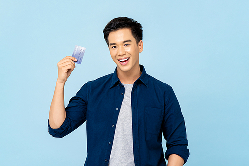 Waist up portrait of happy smiling young handsome Asian man holding credit card in isolated light blue background
