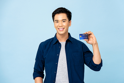 Happy smiling young handsome Asian man holding credit card in isolated light blue background