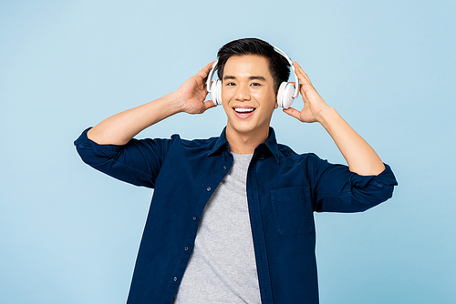 Happy smiling young handsome Asian man listening to music with wireless headphones isolated on light blue studio background